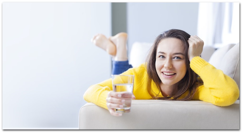 Unge kvinde med gul bluse ligger i sofa og drikker vand
