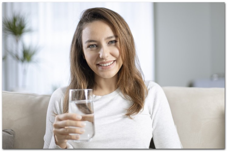 Unge kvinde med hvid bluse sidder i sofa og drikker vand